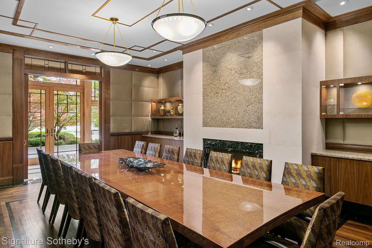 This is an interior shot of a formal dining room, featuring a long, glossy wooden table surrounded by upholstered chairs. A fireplace with a stone surround adds a focal point, while built-in cabinetry and shelving provide storage and display space. The room is well-lit with pendant lighting and recessed lights, creating an elegant and inviting atmosphere.