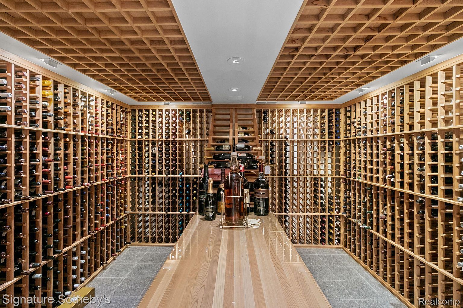 This is an impressive wine cellar featuring extensive wooden racking that houses numerous bottles of wine. A large wooden table sits in the center of the room, displaying several bottles, suggesting a tasting area. The ceiling is also covered in a wooden grid pattern, adding to the cellar's sophisticated and organized aesthetic.