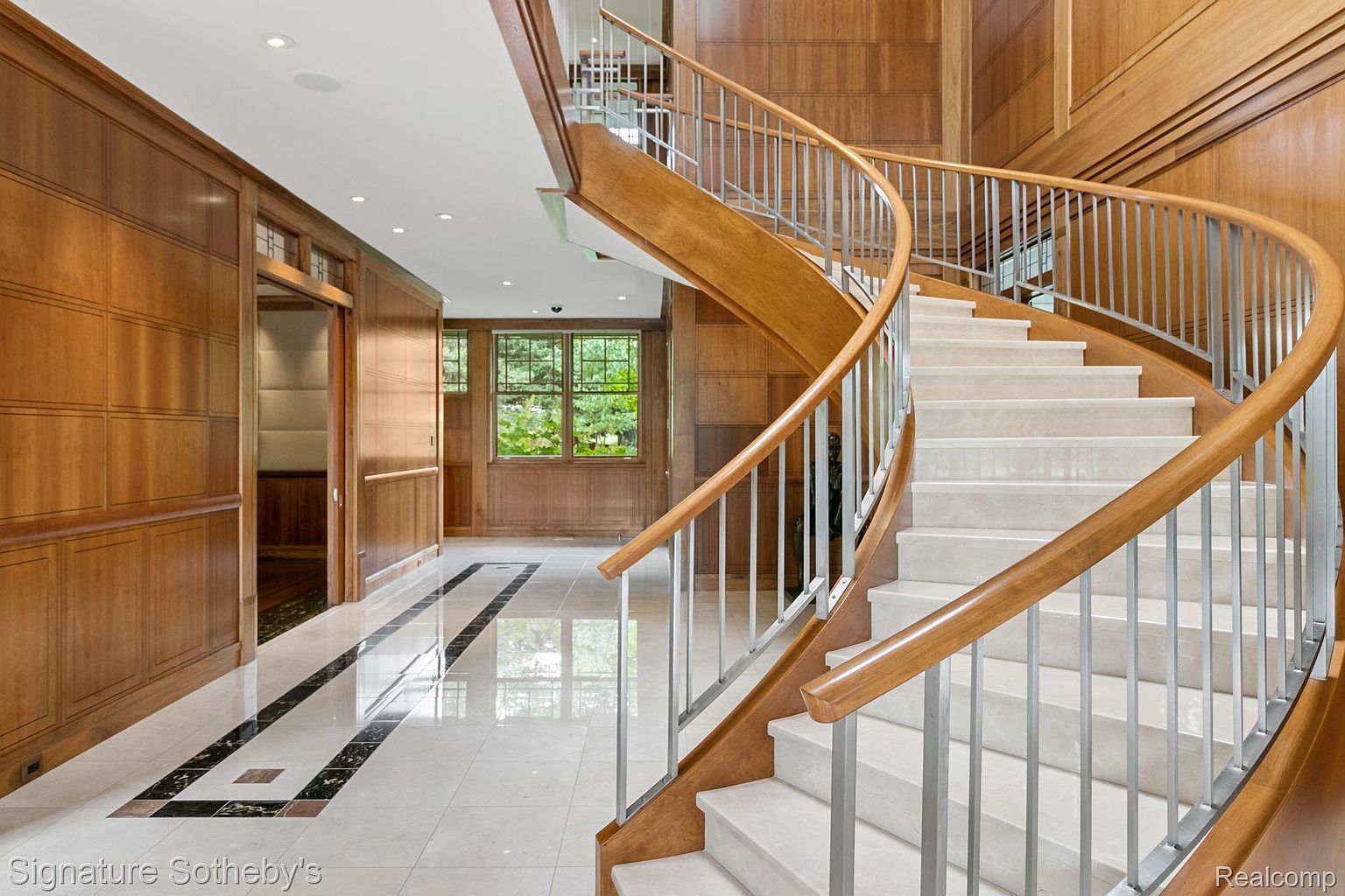 This interior shot showcases a grand hallway featuring a curved staircase with wooden handrails and metal balusters. The walls are paneled with wood, and the flooring is a combination of light tile with dark marble accents. Natural light streams in through a window, highlighting the elegant design and spaciousness of the area.
