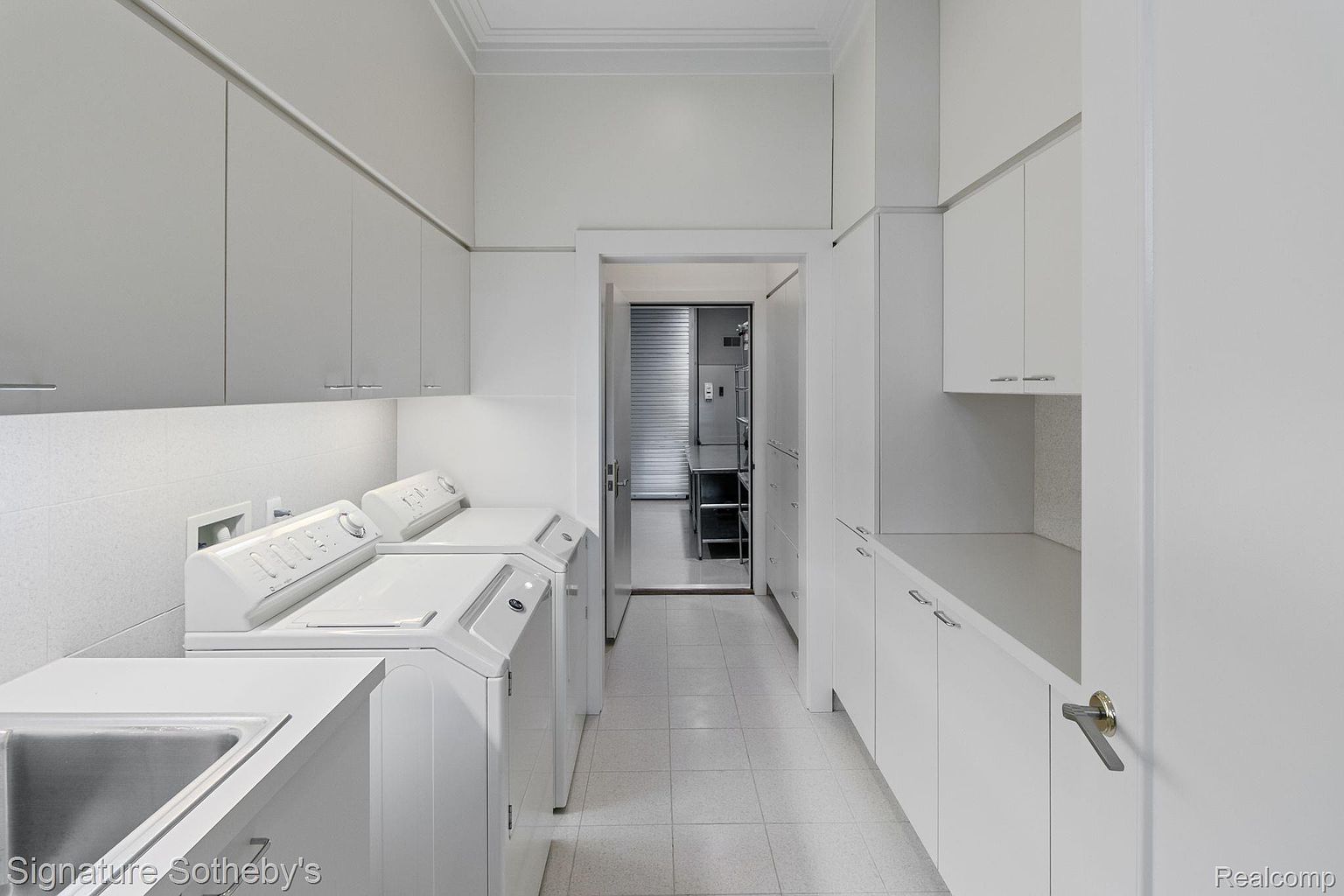This is a well-lit laundry room featuring white cabinetry, a sink, and a washer and dryer set. The room has a clean and organized appearance, with a doorway leading to another room. The overall impression is one of functionality and tidiness.