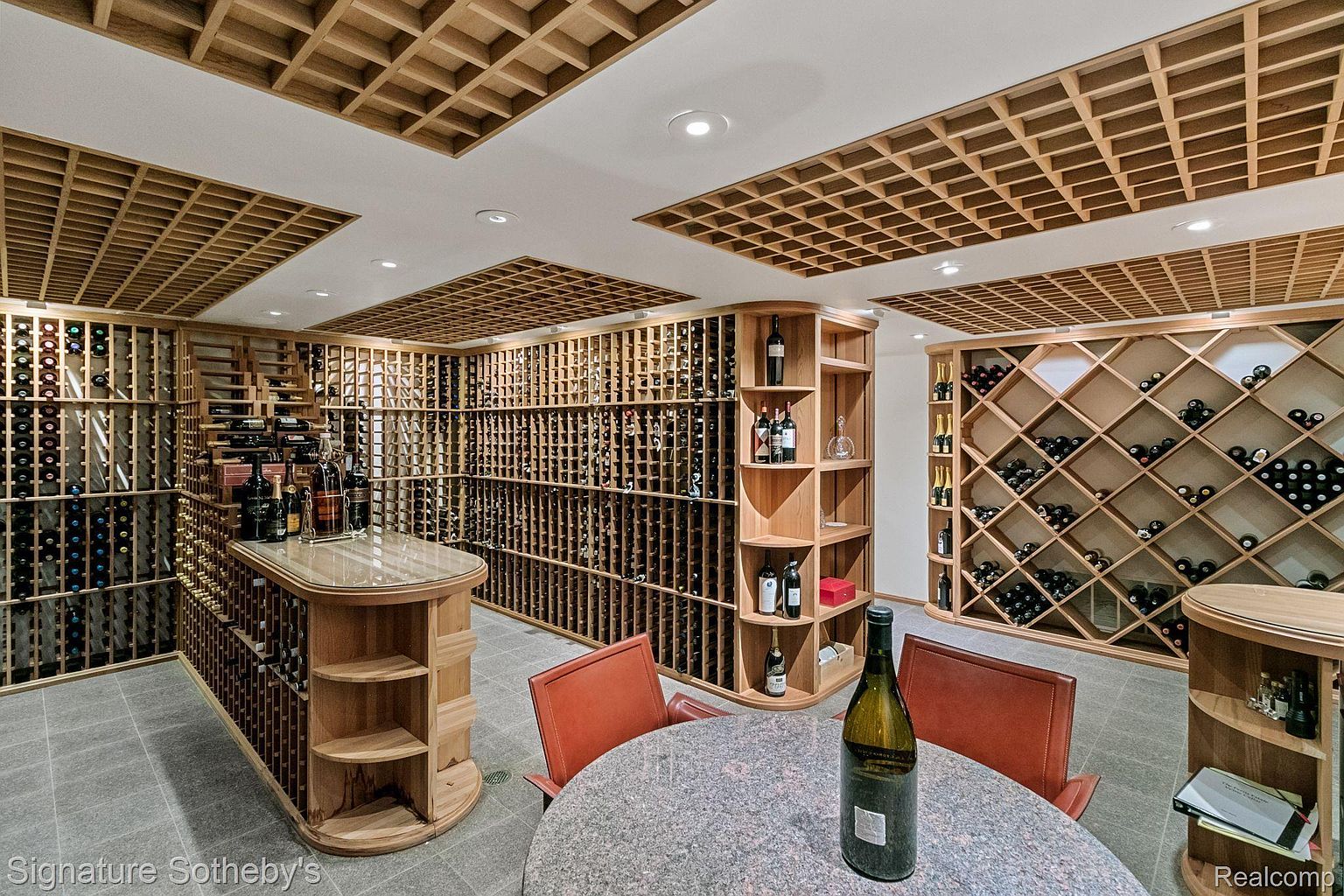 This is a well-appointed wine cellar featuring extensive wooden racking that houses numerous bottles of wine. A round table with chairs sits in the center, suggesting a tasting area. The ceiling has a decorative wooden grid pattern, and the overall impression is one of sophistication and luxury.