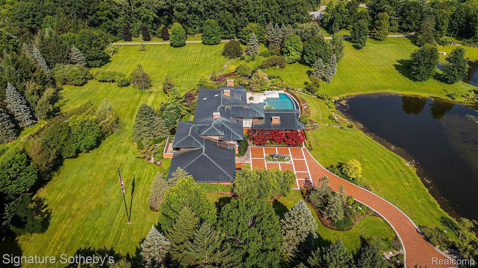 This aerial shot showcases a sprawling estate with a large, modern home featuring a dark roof, a swimming pool, and a pond. The property is surrounded by lush green lawns and mature trees, creating a private and serene setting. A brick driveway leads to the house, adding a touch of elegance.