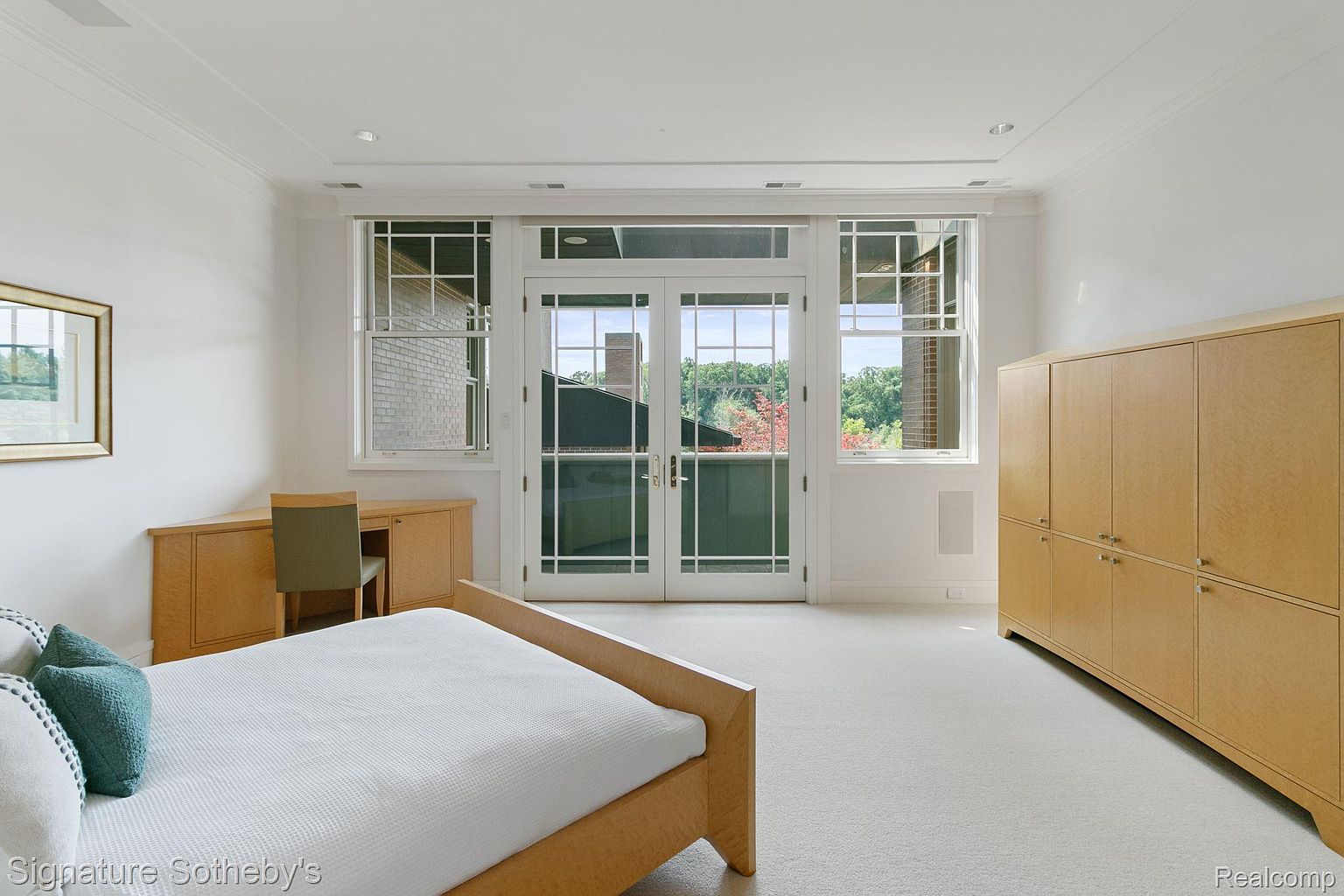 This is a bright and spacious primary bedroom featuring a light wood bed frame with a white mattress and teal pillows. The room has a built-in desk and a large wardrobe, both in a matching light wood finish. Large windows and a set of French doors provide ample natural light and views of the outdoors, enhancing the room's airy and inviting atmosphere.