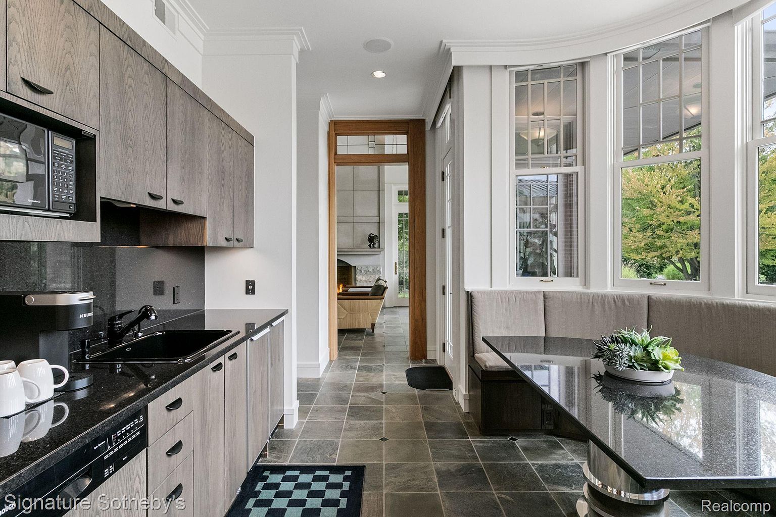 This is a modern kitchen with sleek, gray-toned cabinetry and black countertops. A built-in dining nook with a dark table sits adjacent to large windows, offering natural light. The flooring is a dark tile, and the space opens into another room, creating an open and inviting atmosphere.