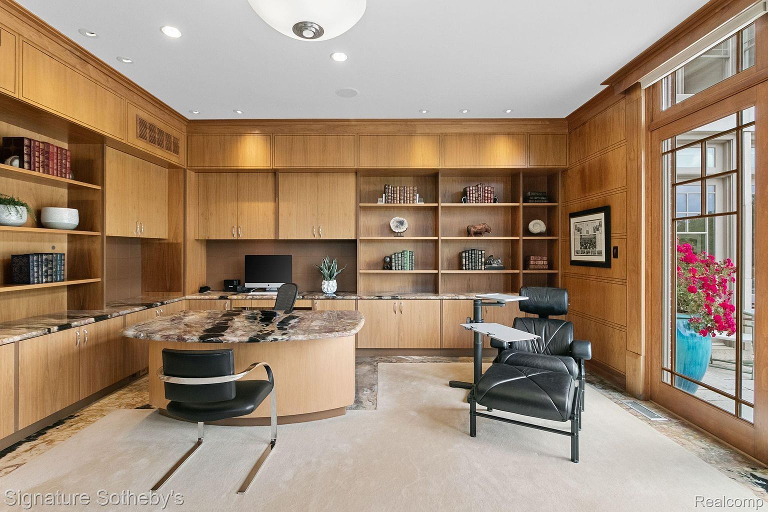 This is an interior shot of a well-appointed office or study. The room features custom wood cabinetry and shelving, a curved desk with a marble countertop, and a comfortable leather chair with a side table. A large window provides natural light and a view of the exterior, enhancing the room's sophisticated and functional design.