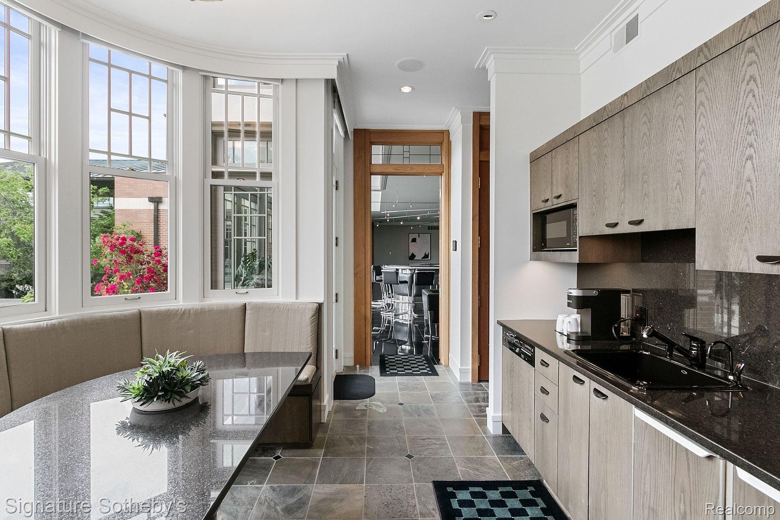 This interior shot showcases a modern kitchen with sleek, light wood cabinetry and dark granite countertops. A cozy breakfast nook with a built-in bench is situated by a large window, offering natural light. The kitchen features modern appliances and a stylish tile floor, leading to an open doorway into another room.
