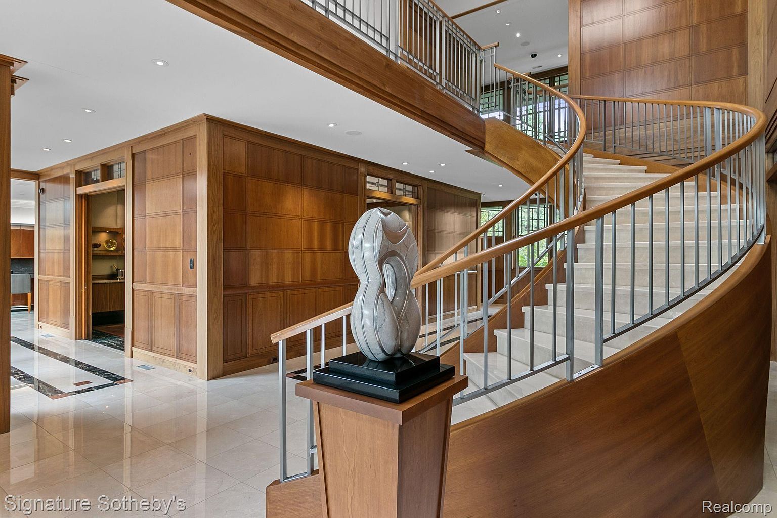 This interior shot showcases a grand hallway featuring a curved staircase with wooden handrails and metal balusters. The walls are paneled with rich wood, and a modern sculpture sits prominently at the base of the stairs. The flooring is polished tile, reflecting the light and enhancing the spacious feel of the entryway.