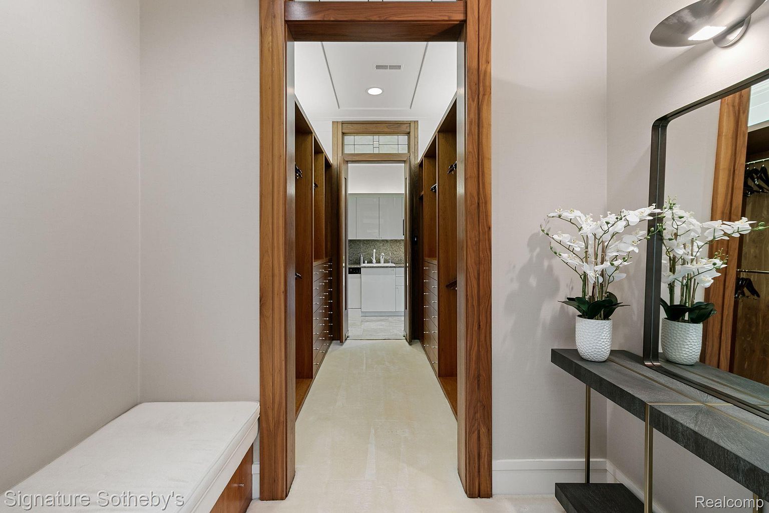 This image showcases a luxurious walk-in closet with custom wooden cabinetry and ample storage space. The closet leads to a bright, modern bathroom, creating a seamless transition between dressing and grooming areas. A stylish console table with floral arrangement adds a touch of elegance to the space.