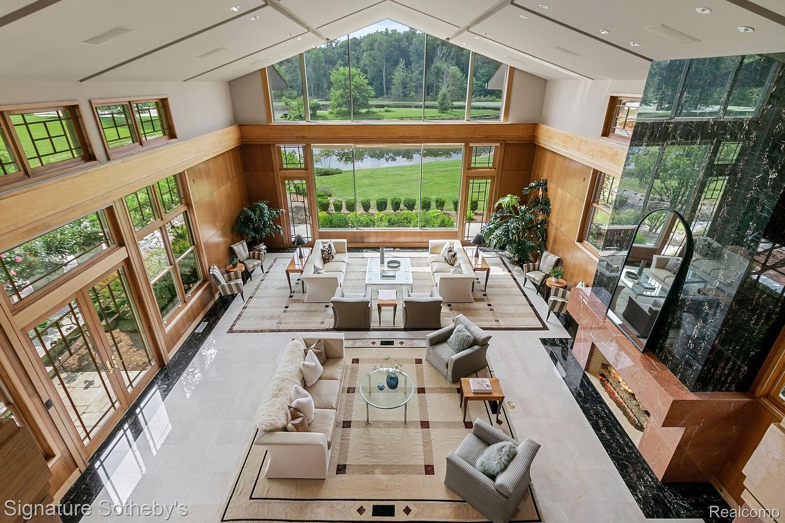 This is a high-angle shot of a grand living room, showcasing a luxurious and spacious interior. The room features large windows offering views of the surrounding landscape, multiple seating areas with plush sofas and armchairs, and a striking fireplace with a marble surround. The overall impression is one of elegance and sophistication, perfect for entertaining or relaxing.