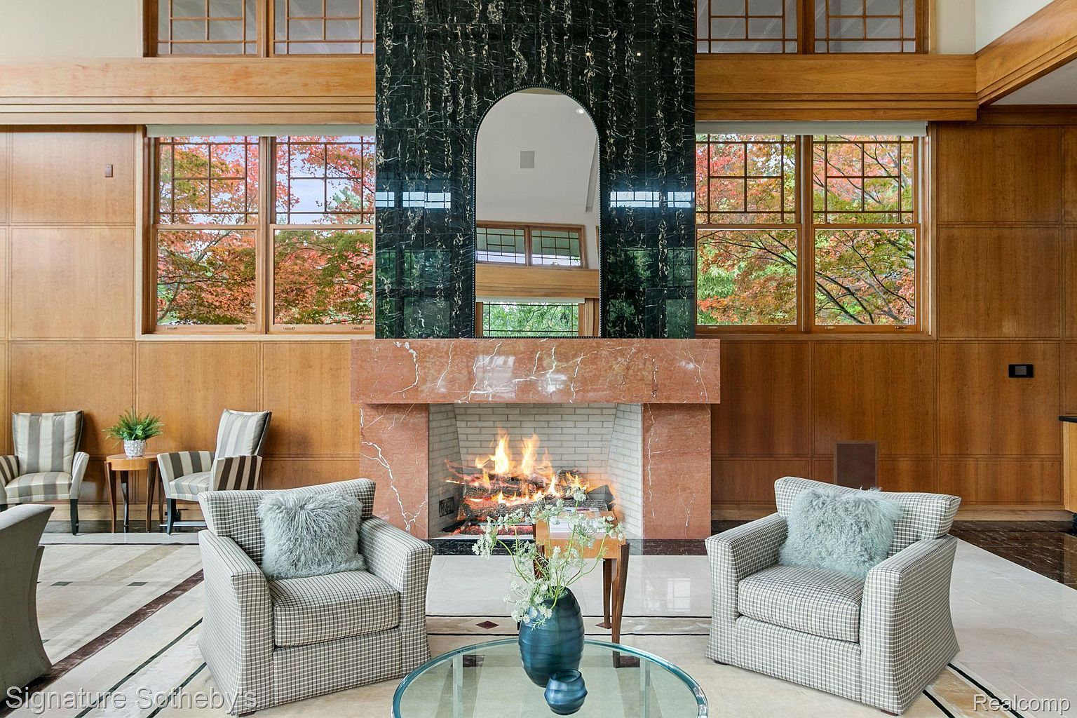 This is an interior shot of a luxurious living room, featuring a grand fireplace with a marble surround and a dark green marble accent above. Two comfortable armchairs flank the fireplace, creating a cozy seating area, while large windows offer views of the colorful autumn foliage outside. The room is paneled with wood and has a patterned marble floor, contributing to an overall elegant and inviting atmosphere.