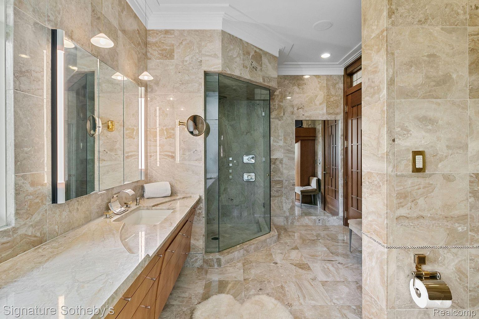 This is a luxurious primary bathroom featuring marble tiling throughout, a glass-enclosed shower, and a vanity with a marble countertop and wooden cabinets. The bathroom also includes a large mirror with integrated lighting and a separate area with a wooden door, creating a spa-like atmosphere. The overall impression is one of elegance and high-end design.