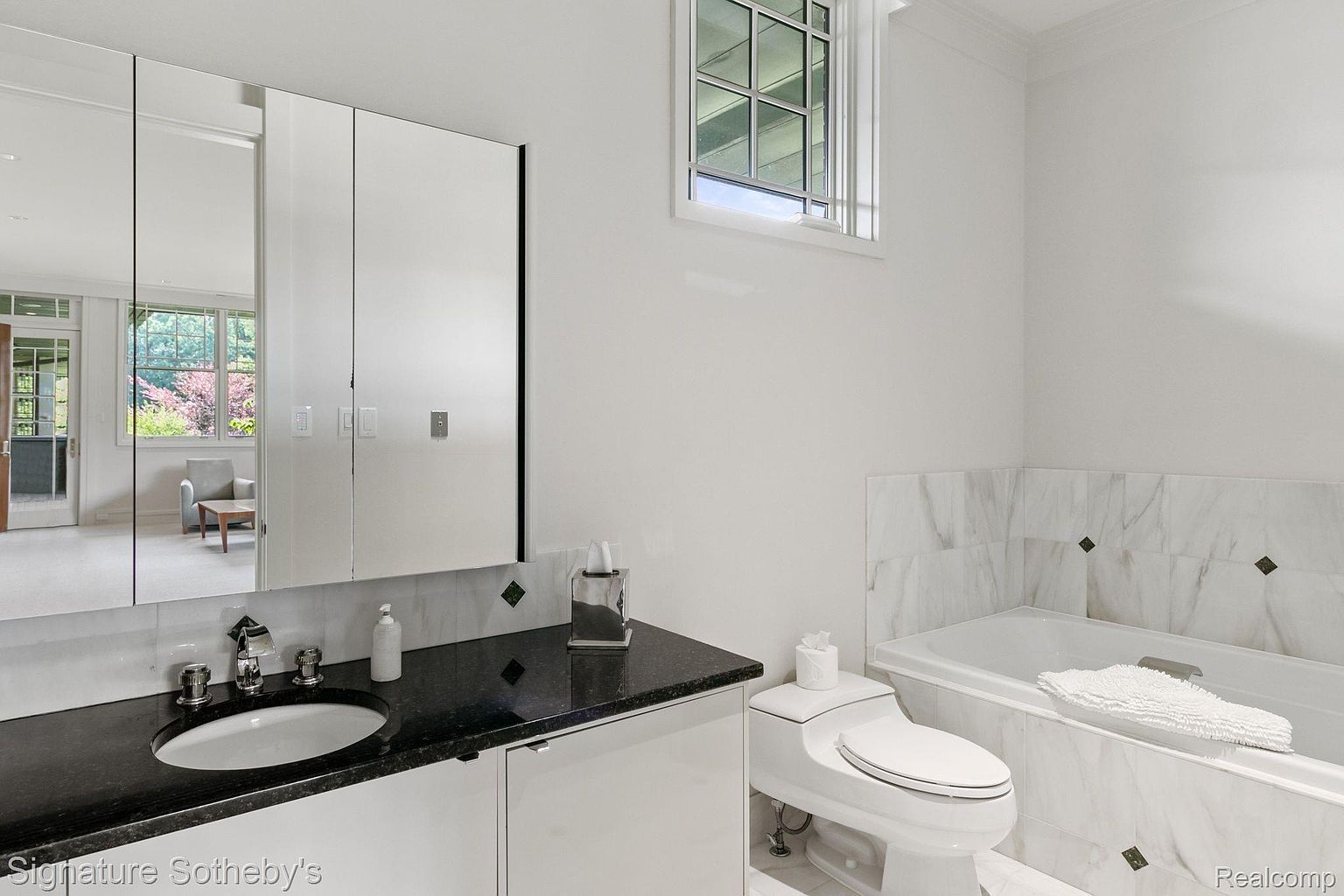 This is a well-lit bathroom featuring a black granite countertop with a white sink and chrome fixtures. A large mirror cabinet hangs above the counter, reflecting a view into another room. The bathroom also includes a toilet and a bathtub with marble tiling, creating a clean and luxurious feel.