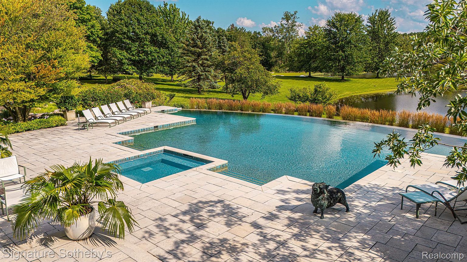 This image showcases a luxurious outdoor pool and spa area. The pool features an infinity edge that blends seamlessly with the surrounding landscape, including a serene lake and lush greenery. The patio is furnished with lounge chairs and a decorative statue, creating an inviting and relaxing atmosphere.