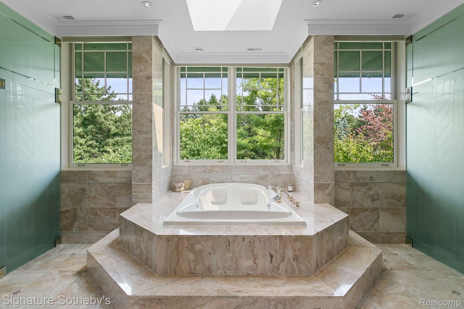 This is a luxurious primary bathroom featuring a large, elevated soaking tub surrounded by marble tiling. Natural light floods the space through multiple windows, offering views of the surrounding greenery. Glass-enclosed shower areas flank the tub, enhancing the spa-like atmosphere.