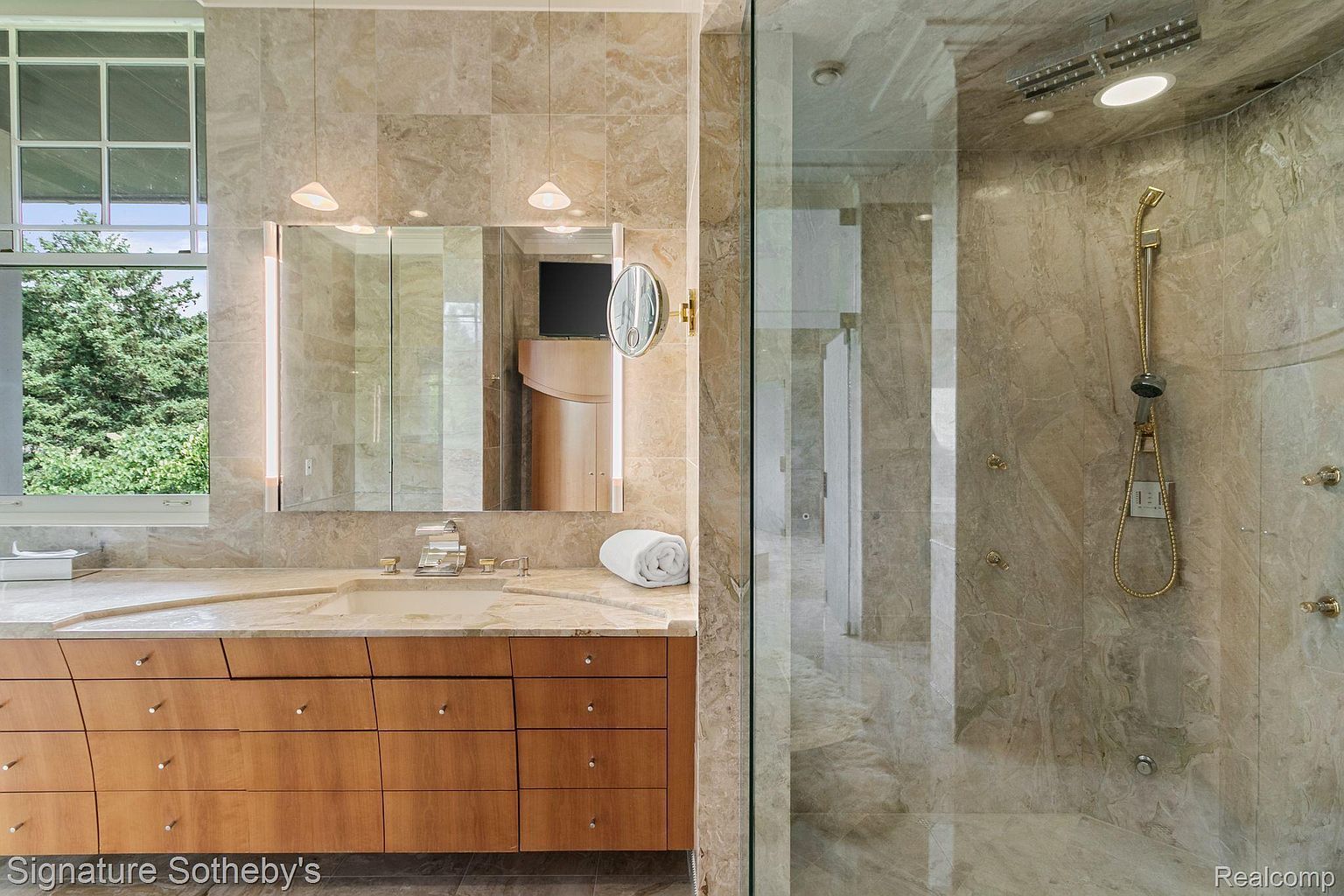 This is a primary bathroom featuring a modern design with marble walls and countertops. The vanity has wood drawers and a built-in sink. A glass-enclosed shower with gold fixtures is visible to the right, and a large window provides natural light.