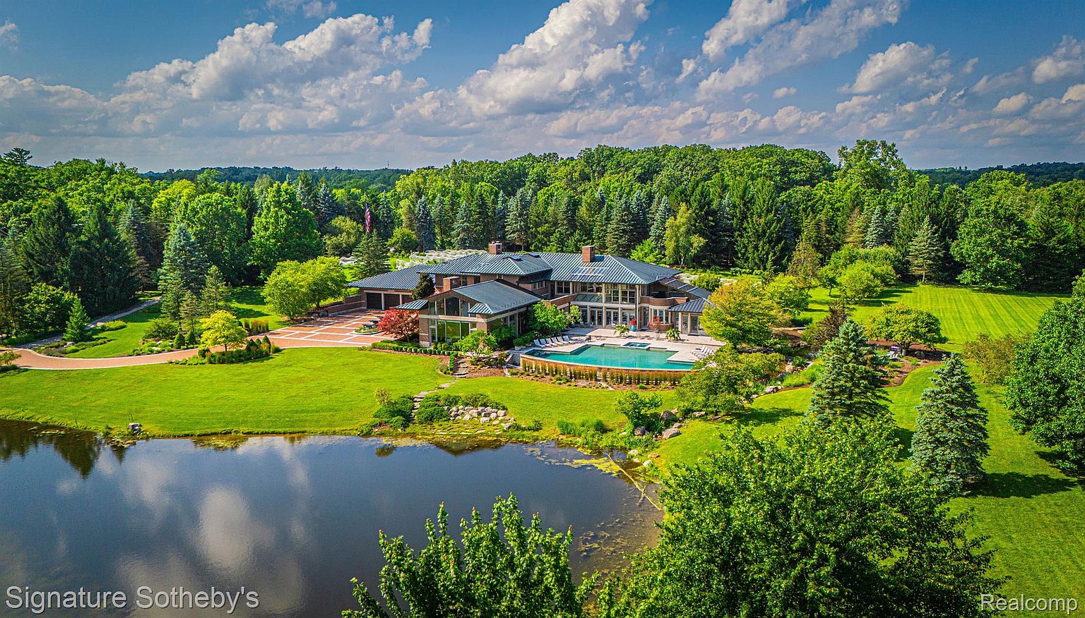 This aerial view showcases a luxurious estate with a sprawling lawn, mature trees, and a serene pond reflecting the sky. The residence features a large pool, a brick driveway, and a sophisticated architectural design with multiple wings and a dark roof. The overall impression is one of grandeur and privacy.