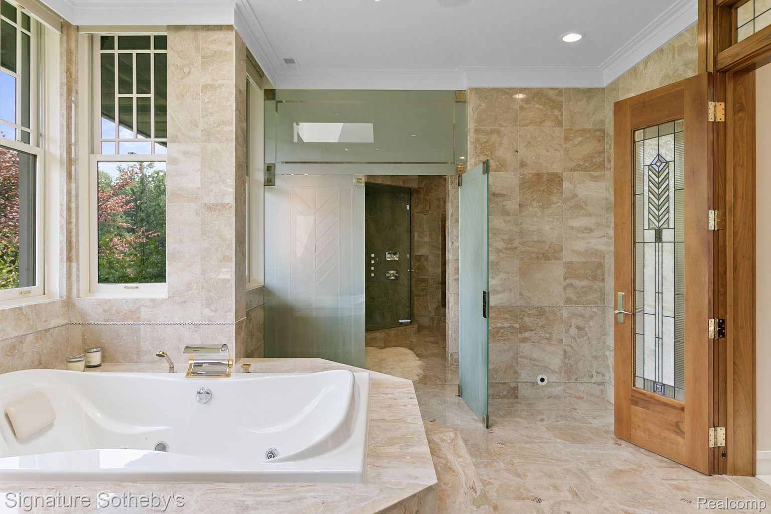 This is a luxurious primary bathroom featuring a large jacuzzi tub with gold fixtures, set against a backdrop of beige marble tiling. A glass-enclosed shower area and a decorative wooden door with stained glass accents add to the room's elegance. The space is well-lit and exudes a spa-like atmosphere.