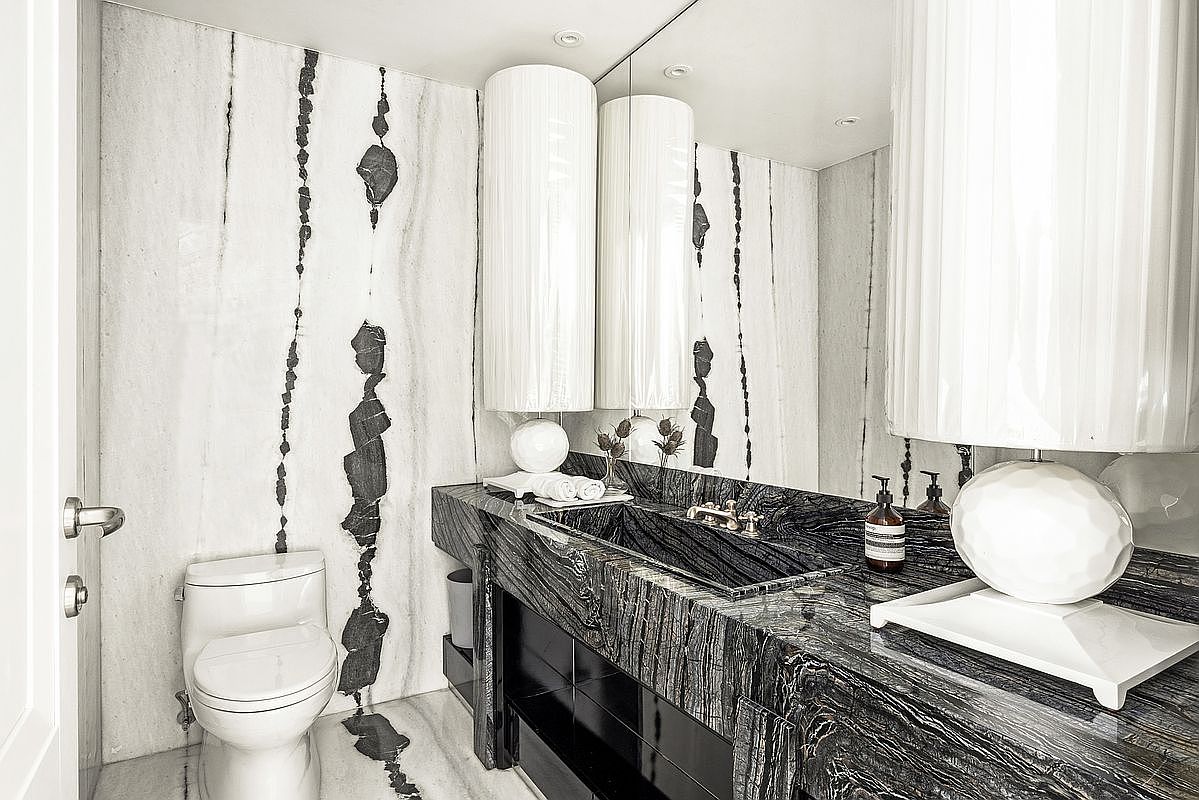 This sophisticated powder room features dramatic, high-contrast stone surfaces with bold, dark veining that extends from the vanity to the wall cladding. A sleek, integrated stone sink sits atop a dark, polished vanity, complemented by a large mirror that enhances the sense of space. The room is accented by a modern, cylindrical lamp and a minimalist toilet, creating a luxurious and moody aesthetic.