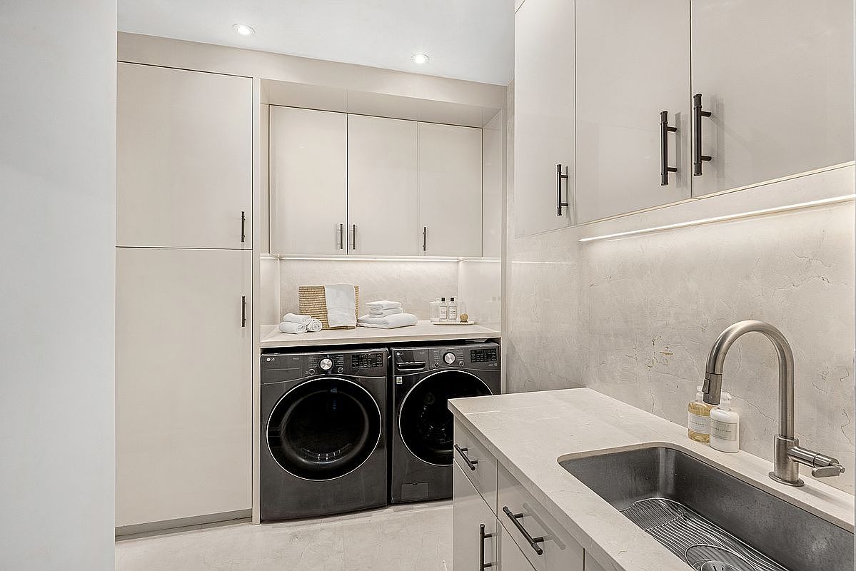 This modern, high-end laundry room features sleek, light-toned cabinetry and a stone countertop with an integrated sink. The space is equipped with dark-toned front-loading washer and dryer units, complemented by under-cabinet lighting and a neutral, minimalist aesthetic. The perspective is a straight-on view that highlights the efficient layout and clean, organized design of the utility space.