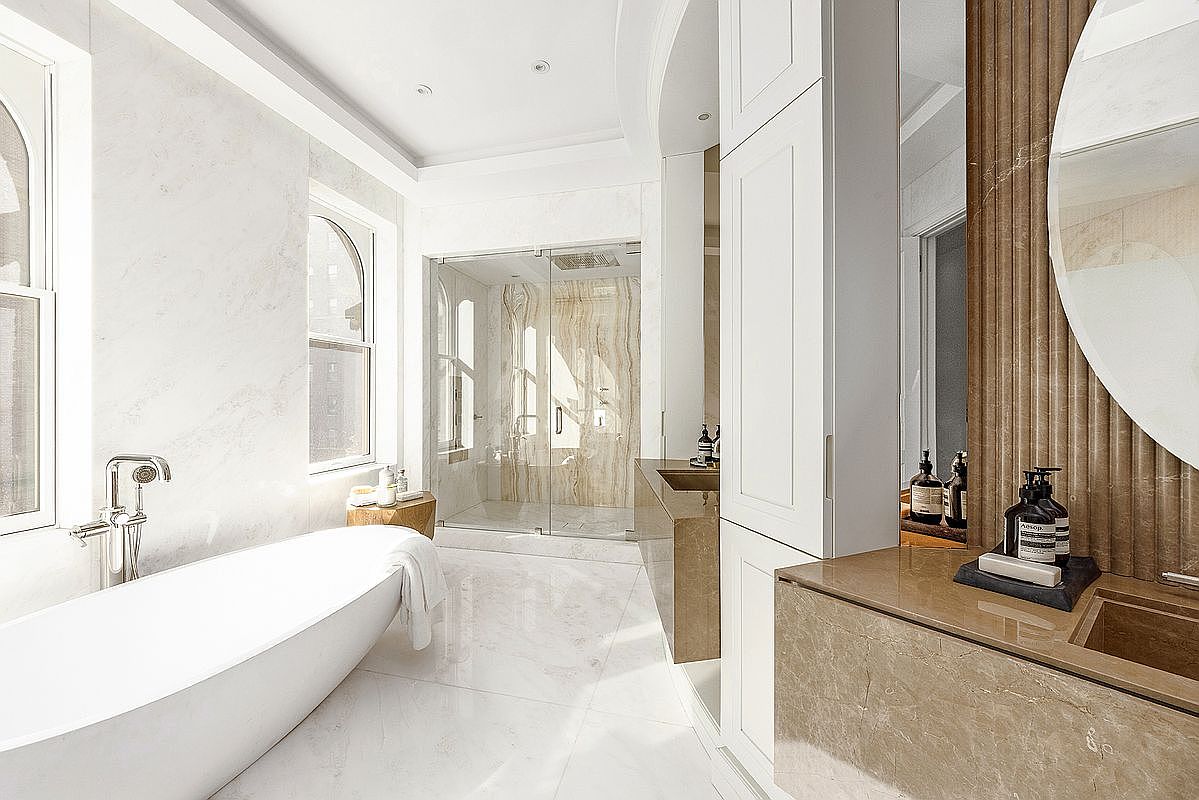 This luxurious primary bathroom features a sleek, freestanding soaking tub positioned near large windows that flood the space with natural light. The room is anchored by a custom stone-topped vanity with a built-in sink and a large circular mirror, complemented by a spacious walk-in shower with glass doors and a textured stone wall. The clean, minimalist aesthetic is enhanced by light-toned flooring and high-end fixtures, creating a serene and sophisticated spa-like atmosphere.
