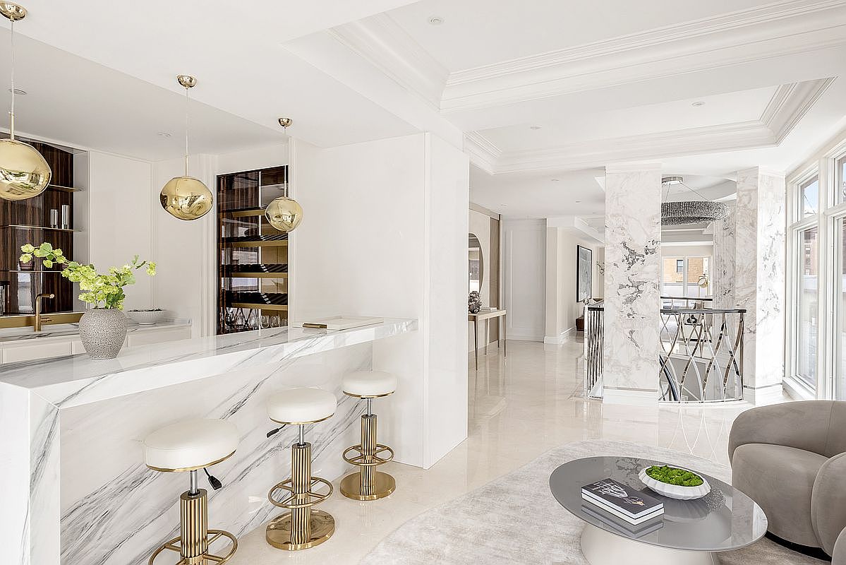 This sophisticated kitchen area features a sleek stone-topped bar with three gold-accented stools, illuminated by modern spherical pendant lights. The space is characterized by a bright, open-concept design with polished light-colored flooring and a built-in wine storage unit. A partial view of a living area with a circular coffee table and a plush armchair is visible in the foreground, while a marble-clad pillar and a staircase railing add architectural interest to the background.