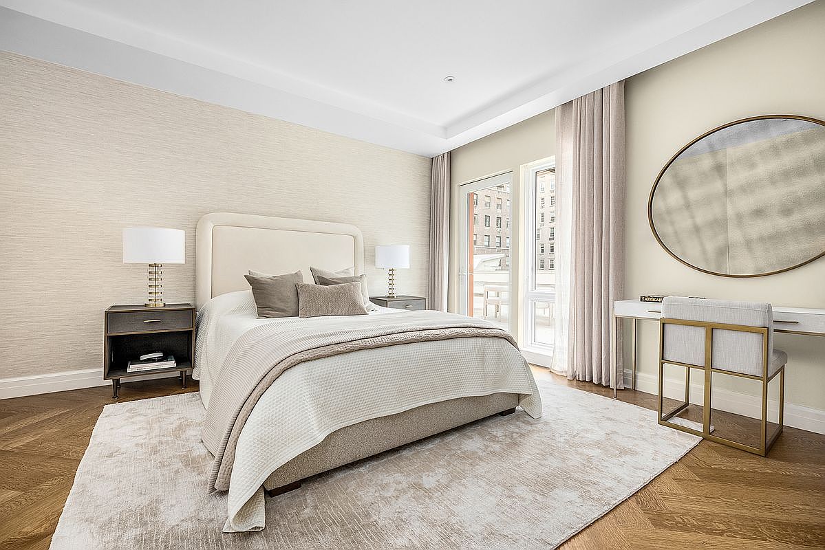 This elegant primary bedroom features a neutral color palette with textured wallpaper and rich herringbone wood flooring. A plush bed with layered linens serves as the focal point, flanked by two dark wood nightstands with matching lamps. To the right, a sleek vanity area with a large circular mirror and a modern chair provides a sophisticated touch, while tall windows with soft drapery allow for ample natural light.