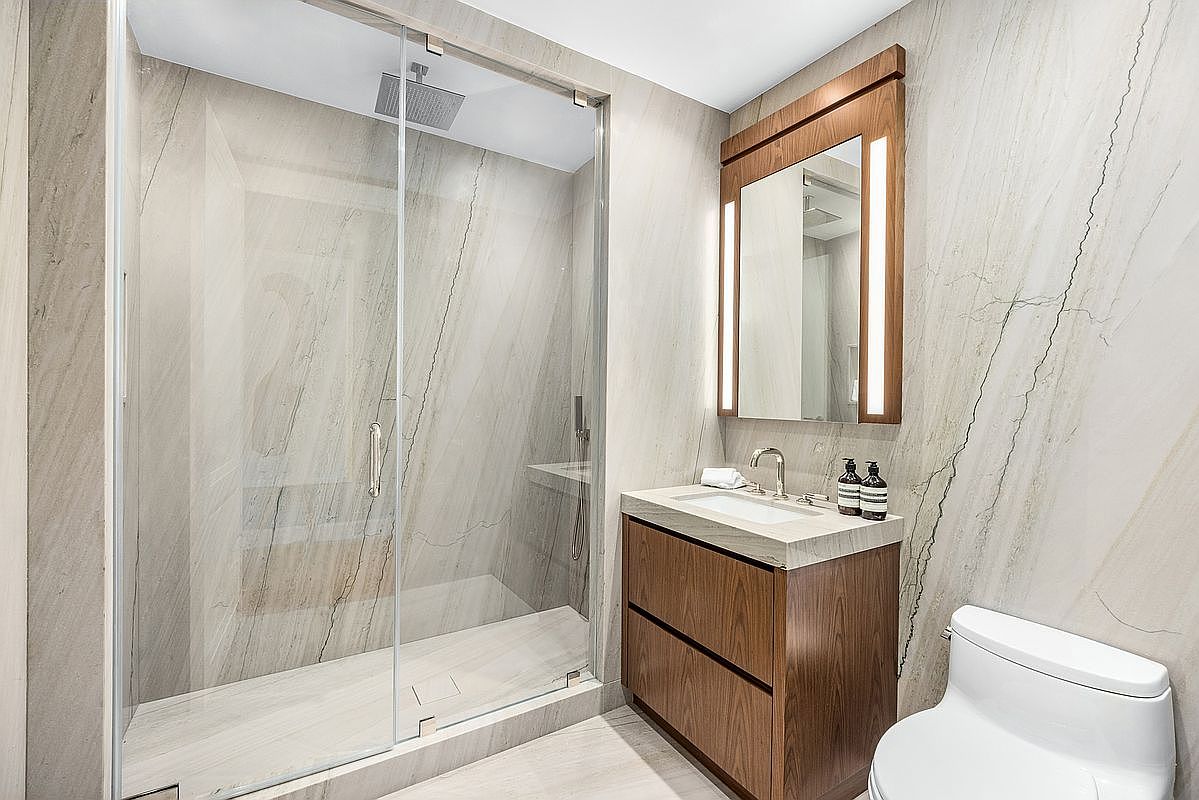 This modern bathroom features a walk-in shower enclosed by a glass panel, showcasing a rainfall showerhead and light-toned stone wall cladding. A floating wood vanity with a stone countertop and a rectangular sink sits beneath a mirror framed in matching wood with integrated vertical lighting. The space is finished with neutral stone flooring and a white toilet, creating a clean, minimalist, and high-end aesthetic.
