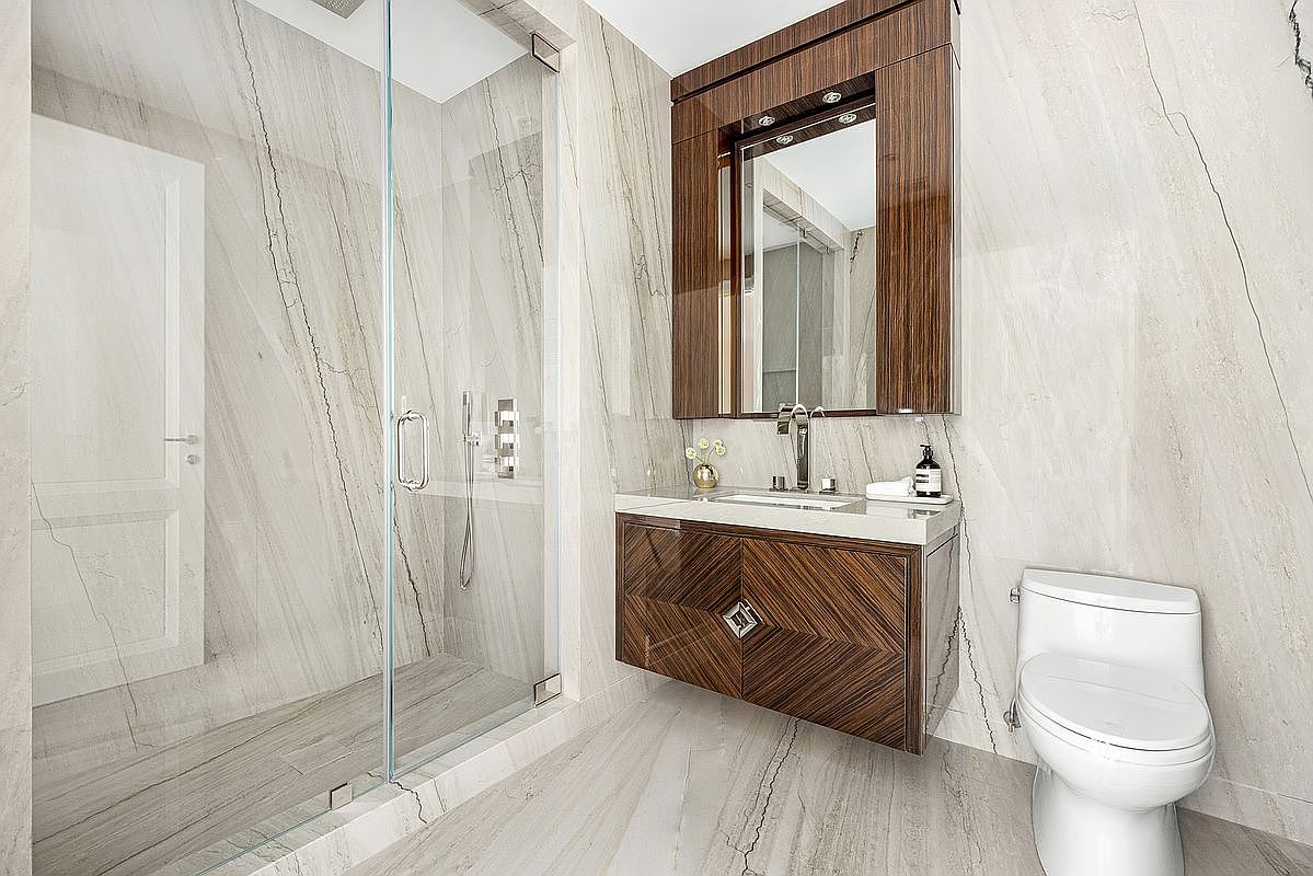 This modern bathroom features large-format stone wall and floor tiles with prominent veining, creating a cohesive and luxurious aesthetic. A floating vanity with a rich, dark wood finish and a geometric pattern serves as the focal point, complemented by a matching framed mirror and a glass-enclosed walk-in shower. The space is completed by a white toilet and minimalist chrome fixtures, offering a clean and sophisticated design.