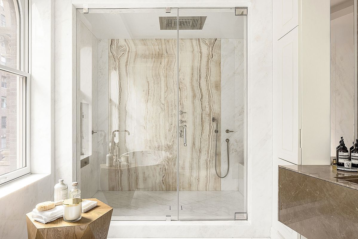 This luxurious primary bathroom features a spacious walk-in shower enclosed by glass panels, showcasing a striking, vertically-veined stone accent wall. The room is finished with light-toned stone surfaces, a sleek vanity area with dark stone countertops, and a geometric wooden side table in the foreground. The bright, airy atmosphere is enhanced by natural light streaming through a large window, creating a sophisticated and serene spa-like environment.