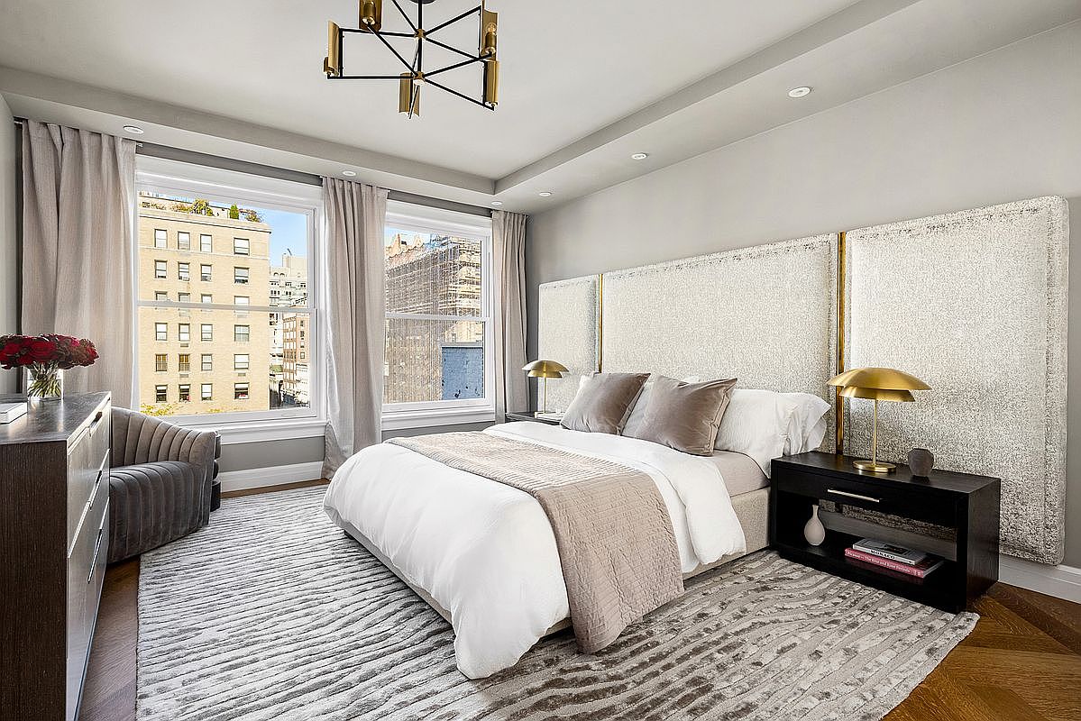 This elegant primary bedroom features a large, textured upholstered headboard that spans the width of the wall, complemented by dark wood nightstands and metallic dome lamps. A plush, patterned area rug sits atop rich herringbone-patterned wood floors, while a cozy armchair and a vase of red roses add a touch of warmth. The room is bathed in natural light from two large windows that offer a view of the surrounding city architecture.