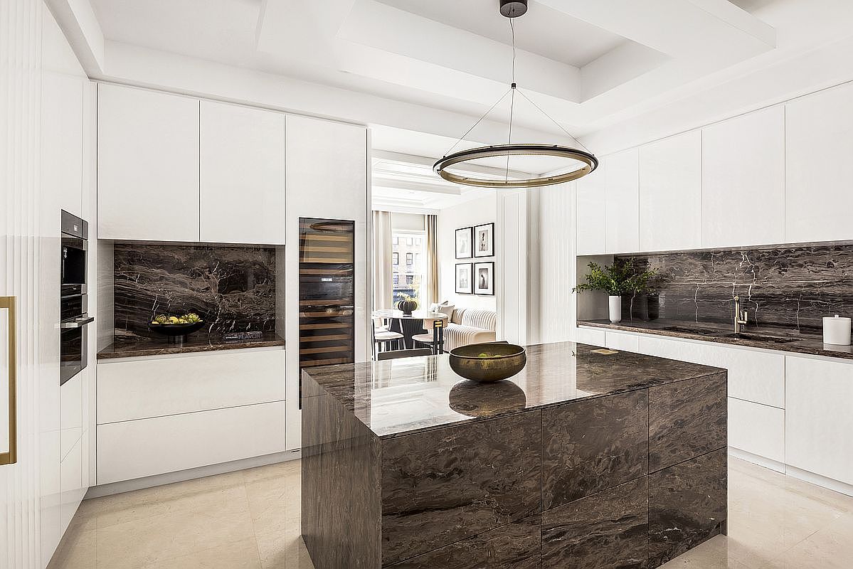 This sophisticated, modern kitchen features high-gloss white cabinetry paired with striking dark stone surfaces on the backsplash and central island. A large, circular pendant light serves as a focal point above the island, while integrated appliances and a wine cooler maintain a seamless, minimalist aesthetic. The perspective looks directly into the kitchen, with a glimpse of an adjacent dining area visible through the open doorway in the background.
