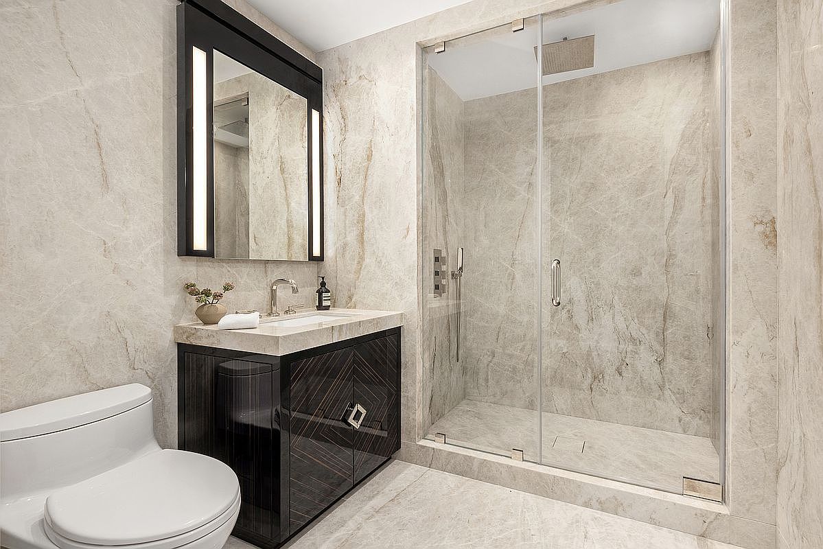 This modern bathroom features light-toned stone wall and floor tiles with subtle veining, creating a cohesive and luxurious aesthetic. A dark, patterned vanity cabinet with a stone countertop and a sleek, illuminated mirror serves as the focal point, complemented by a glass-enclosed walk-in shower. The perspective is a straight-on shot that highlights the clean lines and high-end finishes of the space.