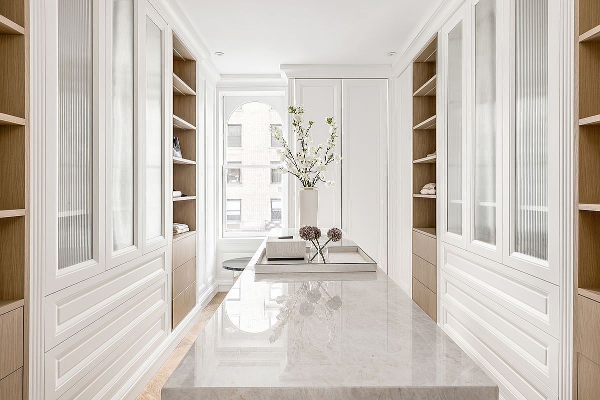 This elegant walk-in closet features a symmetrical layout with white cabinetry, glass-fronted doors, and light wood shelving. A central island with a polished stone countertop serves as a focal point, topped with a decorative vase of flowers and a tray. The space is bright and airy, framed by a window at the end of the room that allows natural light to illuminate the refined, organized storage area.