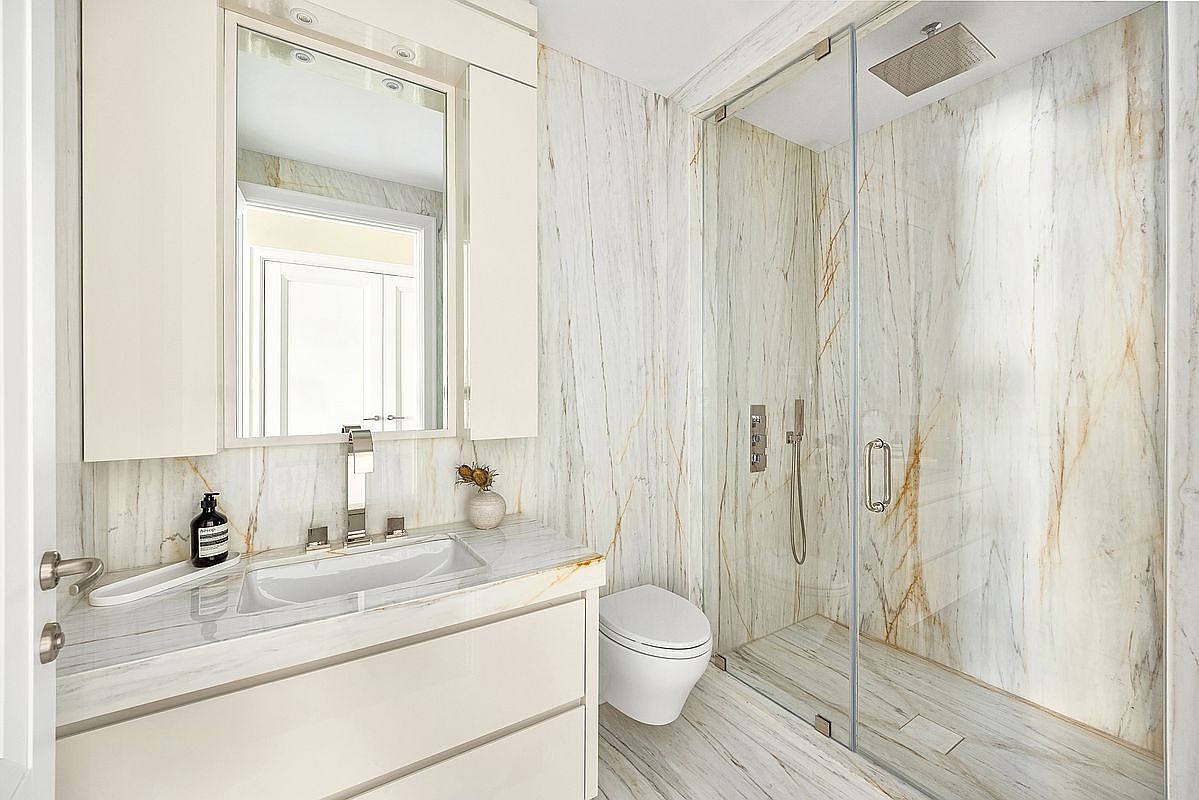 This modern bathroom features elegant, light-toned stone surfaces with prominent veining throughout the walls and vanity countertop. The space includes a sleek, wall-mounted toilet, a contemporary vanity with a rectangular sink, and a walk-in shower enclosed by a glass panel. The perspective is a wide-angle shot that captures the cohesive design and high-end finishes of the room.