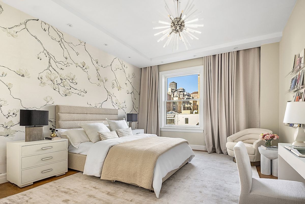 This elegant primary bedroom features a sophisticated neutral color palette and a striking floral-patterned accent wall behind the upholstered headboard. The room is anchored by a plush area rug and illuminated by a modern, starburst-style chandelier, while large windows draped in floor-length curtains offer a view of the city skyline. A dedicated vanity area and a cozy armchair complete the space, creating a serene and luxurious retreat.