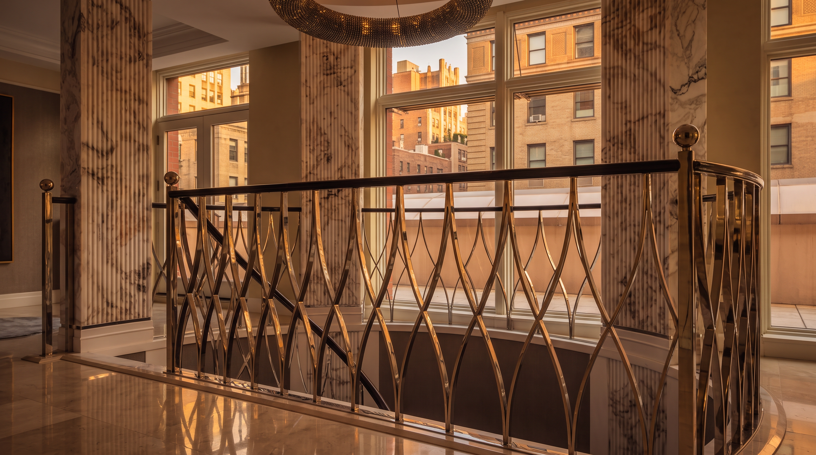 This elegant landing area features a striking circular chandelier suspended above a curved staircase with a polished metal lattice railing. The space is defined by large stone-clad columns and expansive windows that provide ample natural light and views of the surrounding urban architecture. The polished stone flooring reflects the bright, airy atmosphere, creating a sophisticated and luxurious impression.