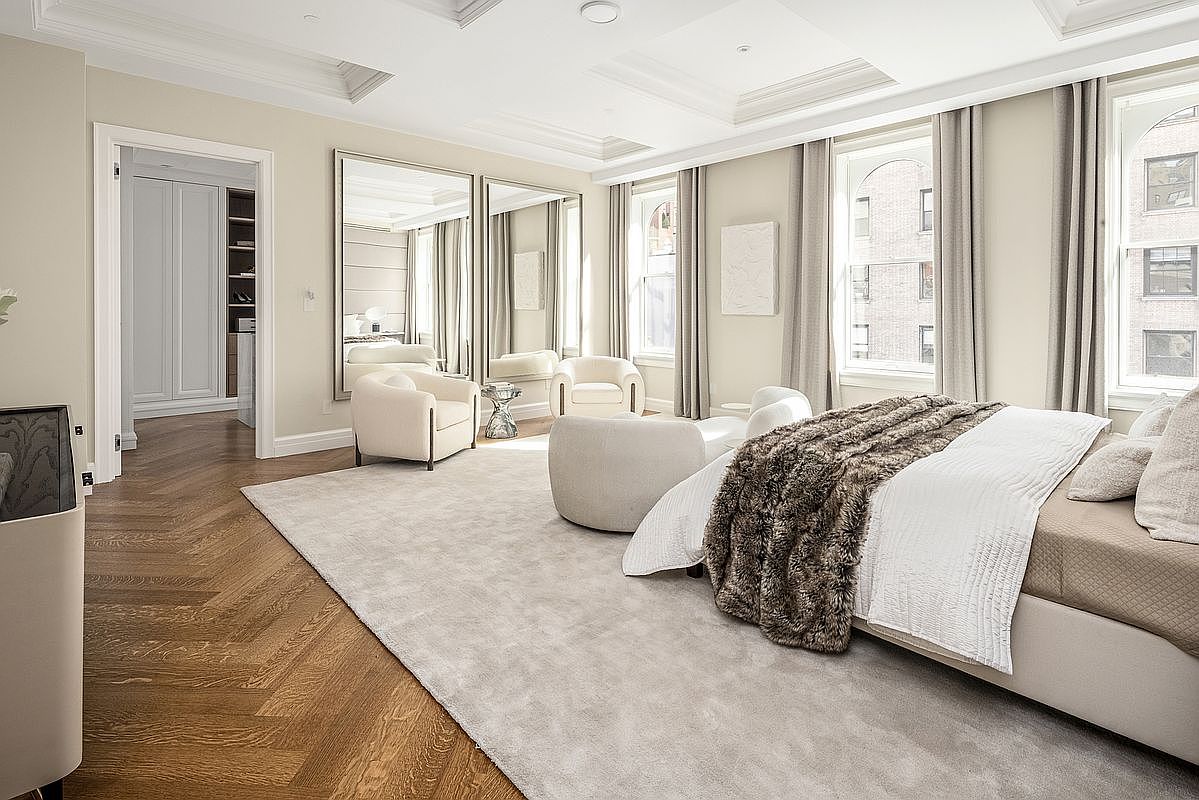 This spacious primary bedroom features elegant neutral tones, herringbone-patterned wood flooring, and a large area rug that anchors the space. The room is furnished with a plush bed topped with a textured throw, two curved accent chairs, and a pair of tall, framed mirrors that enhance the sense of depth. Large windows with floor-length curtains allow natural light to fill the room, creating a serene and sophisticated atmosphere.