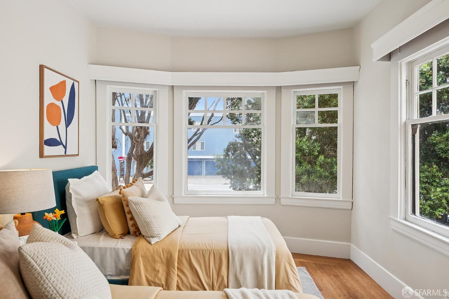 This cozy bedroom features a bed dressed in warm, neutral-toned linens and a teal headboard, accented by decorative pillows. A prominent bay window configuration allows ample natural light to fill the space, offering a view of the outdoors. The room is finished with light-colored walls and warm-toned wood flooring, creating a bright and inviting atmosphere.