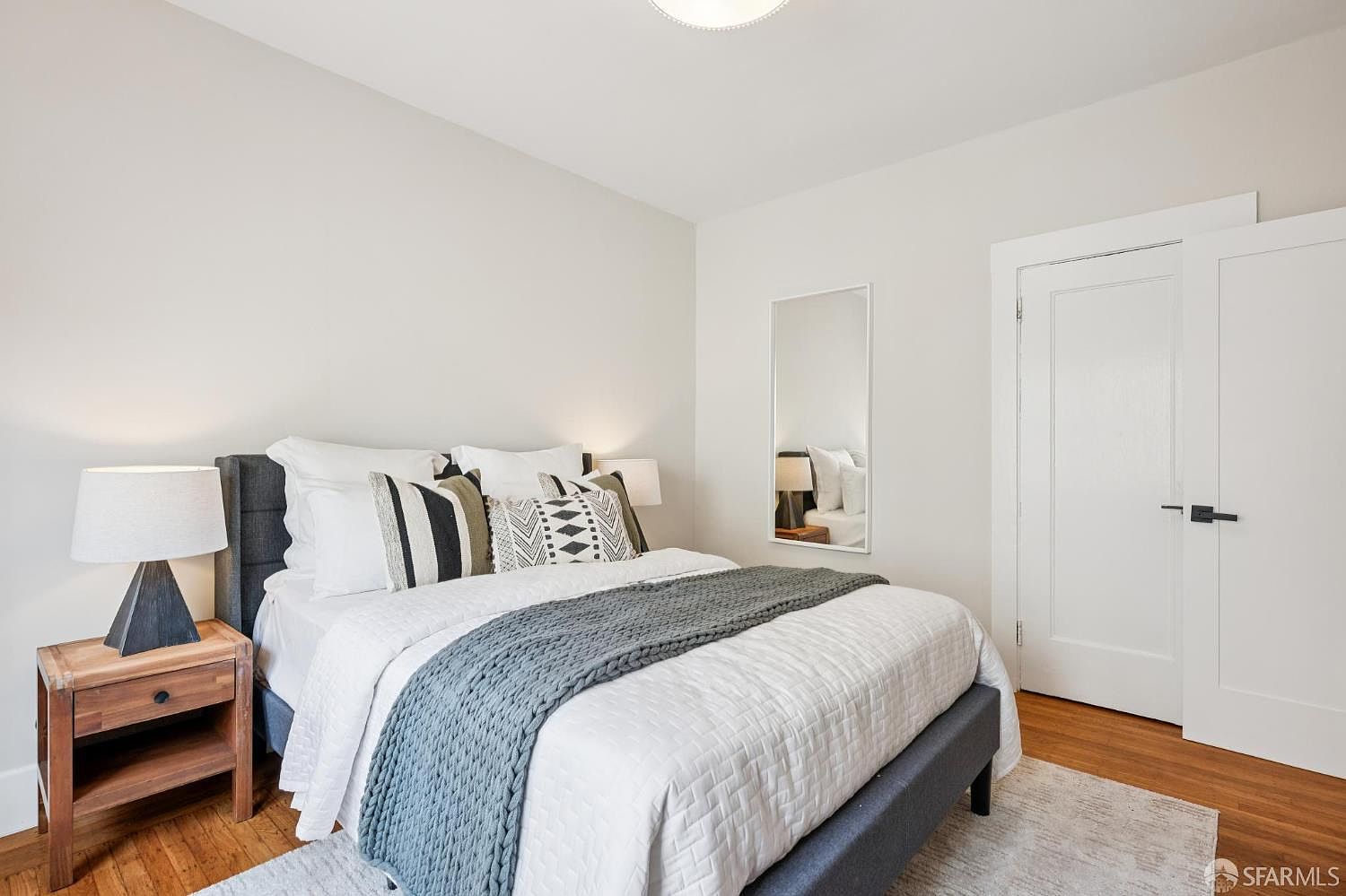 This bright and inviting bedroom features a queen-sized bed with a dark upholstered headboard, layered with white linens and a textured grey throw blanket. A wooden nightstand with a modern lamp sits to the left, while a tall, slim mirror is mounted on the wall, reflecting the room's neutral color palette. The space is finished with warm-toned hardwood flooring and a white door leading to a closet, creating a clean and comfortable atmosphere.