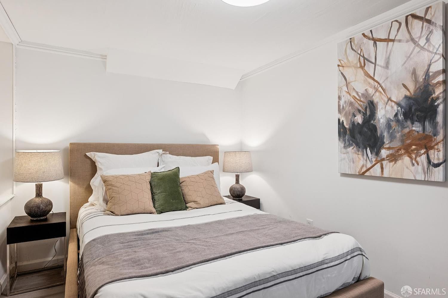 This cozy bedroom features a neutral color palette with a beige upholstered headboard and a bed dressed in white linens with a grey throw blanket. Two matching nightstands with lamps flank the bed, while a large, abstract canvas painting hangs on the wall to the right, adding a touch of artistic flair. The room is brightly lit, creating a clean and inviting atmosphere suitable for a guest space.