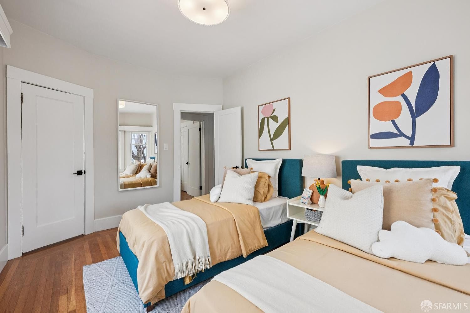 This bright and inviting guest bedroom features two twin beds with vibrant teal upholstered headboards and neutral-toned bedding. The room is decorated with modern abstract wall art, a central light fixture, and a full-length wall mirror that enhances the sense of space. Warm wood flooring and light-colored walls create a clean, contemporary aesthetic suitable for a comfortable living environment.
