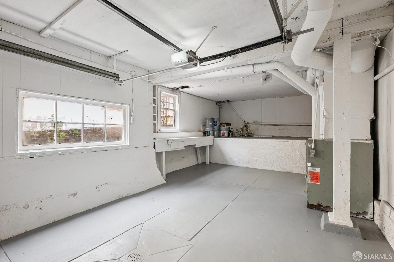 This interior view of a garage features painted white walls, a grey concrete floor, and exposed ceiling pipes. A workbench is positioned against the back wall under a window, and a garage door opener is mounted to the ceiling. The space is utilitarian and functional, showing signs of storage and maintenance.