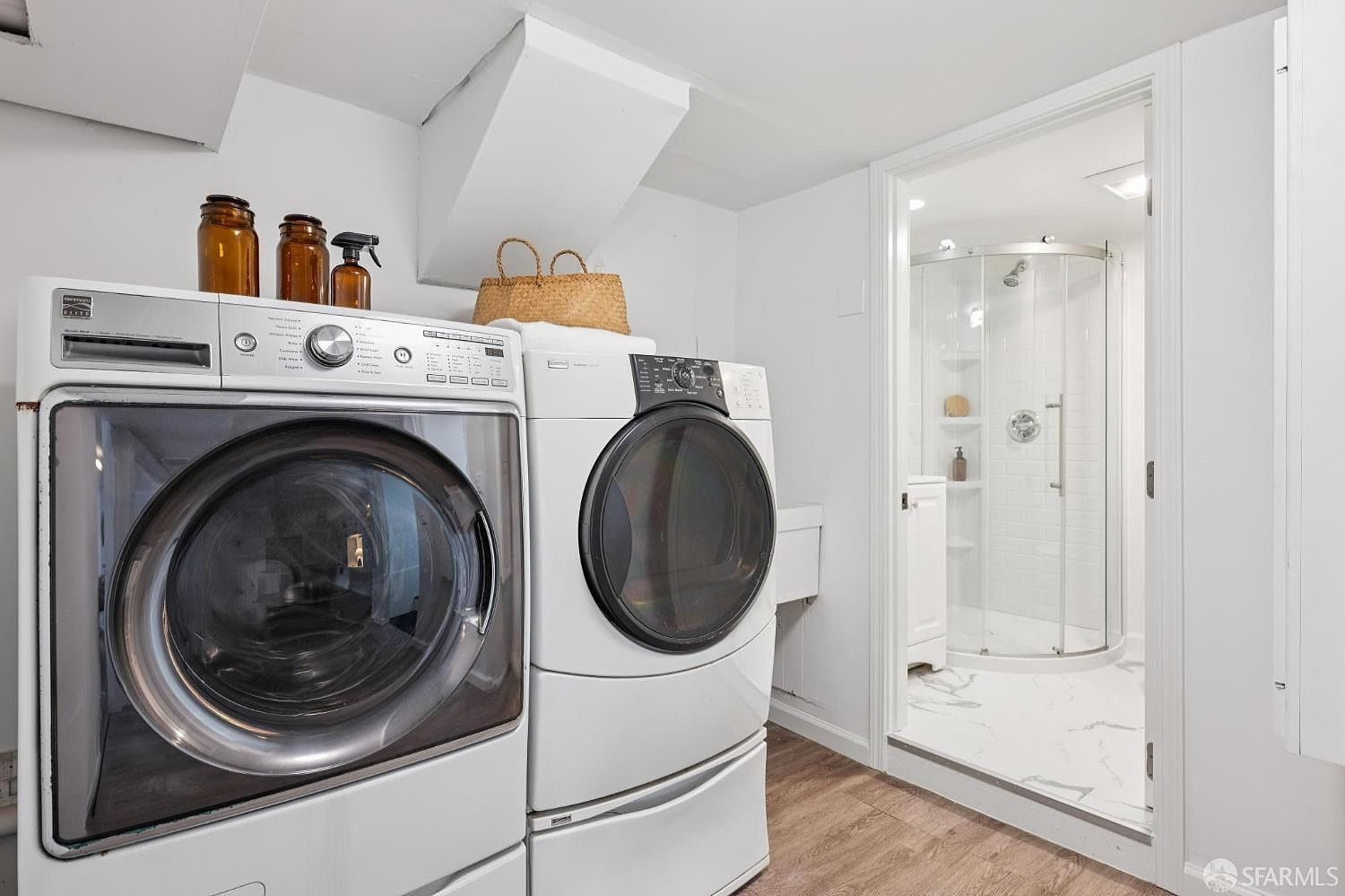 This functional laundry room features a side-by-side washer and dryer set positioned against a white wall. A woven basket sits atop the dryer, while amber-colored glass bottles are arranged on the washer, adding a touch of organization. A doorway leads directly into a bright bathroom with a glass-enclosed shower and patterned tile flooring, creating a convenient and efficient layout.