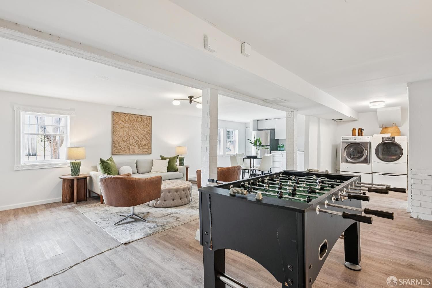This open-concept basement features a versatile layout that combines a cozy living area with a game room and laundry space. The living area is anchored by a neutral sofa, a tufted ottoman, and a leather swivel chair, while a foosball table serves as a prominent focal point in the foreground. The space is finished with light wood-look flooring and white-painted brick columns, creating a bright and functional multi-purpose environment.