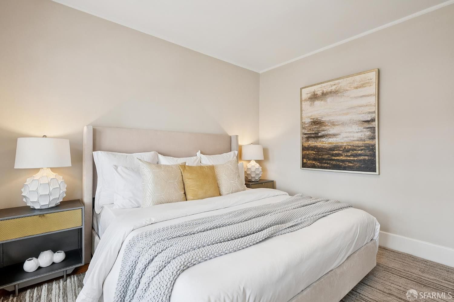 This serene primary bedroom features a neutral color palette and a plush, upholstered headboard as the focal point. The bed is dressed in crisp white linens with a textured gray throw blanket and decorative gold and patterned pillows, flanked by two modern bedside tables with unique sculptural lamps. An abstract piece of wall art hangs above the bed, adding a sophisticated touch to the minimalist, well-lit space.