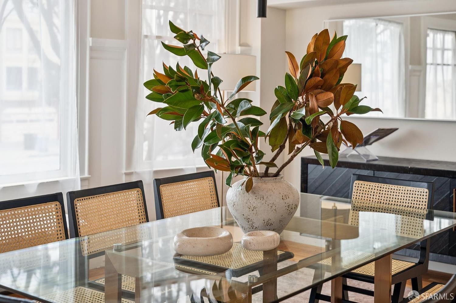 This elegant dining room features a glass-topped table surrounded by chairs with woven cane detailing and dark frames. A large, textured ceramic vase filled with lush magnolia branches serves as a striking centerpiece, complemented by two smaller matching bowls on the table. The space is bright and airy, with large windows draped in sheer curtains and a dark sideboard visible in the background, creating a sophisticated and inviting atmosphere.