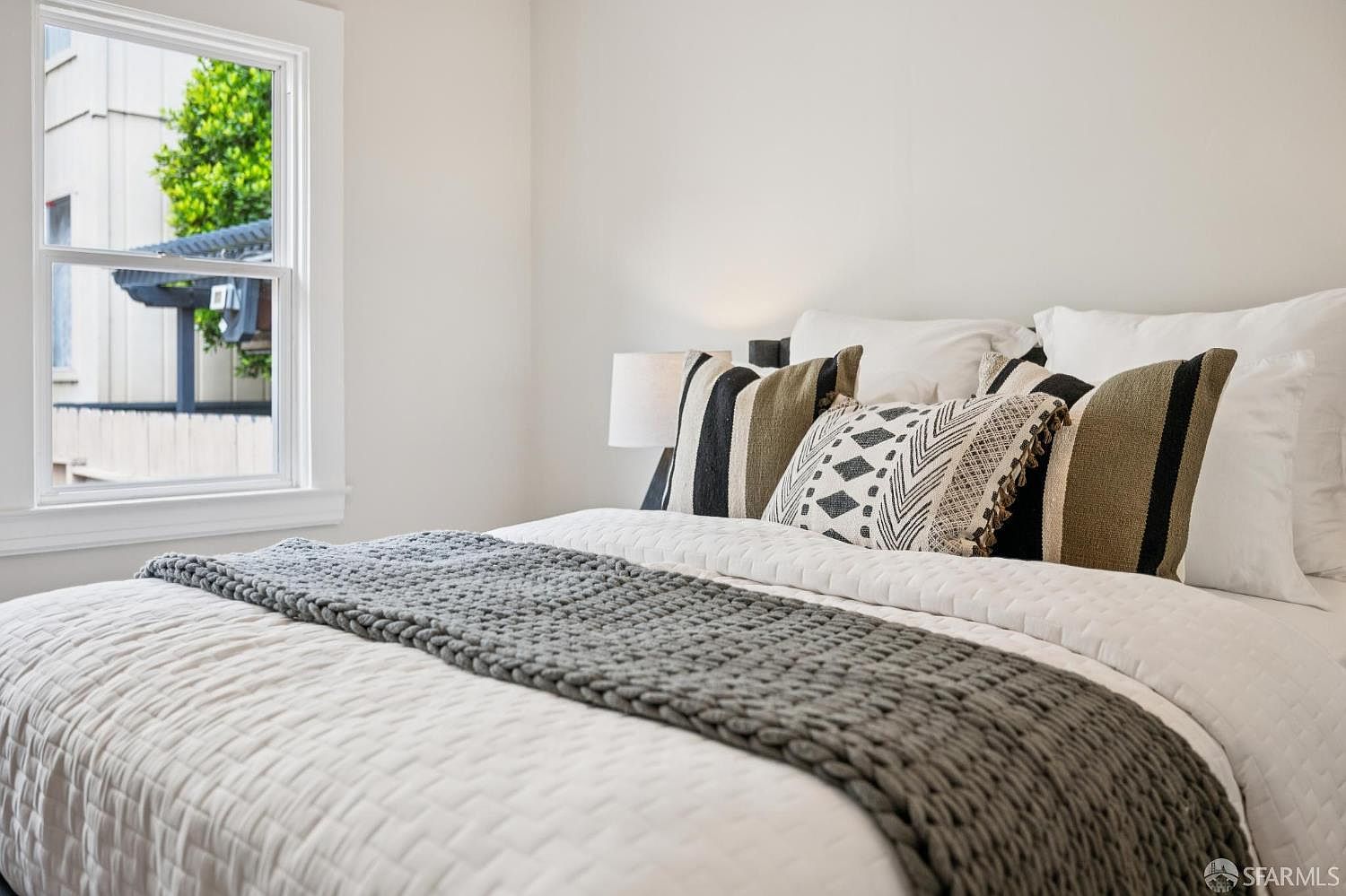 This cozy bedroom features a neatly made bed with a textured white quilt and a thick, dark gray knit throw blanket. The bed is adorned with a collection of decorative pillows in neutral tones and geometric patterns, creating a comfortable and inviting atmosphere. A window to the left allows natural light to brighten the space, offering a view of the exterior surroundings.