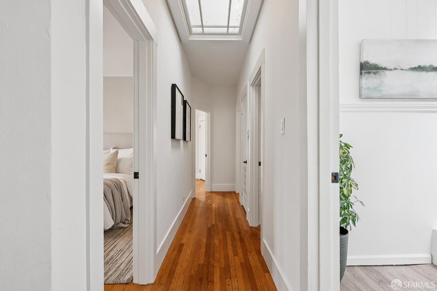 This bright, clean hallway features warm-toned wood flooring and crisp white walls, creating an inviting and airy atmosphere. A skylight overhead provides natural illumination, while multiple doorways lead to various rooms, including a glimpse of a bedroom on the left. The perspective looks down the length of the corridor, highlighting the home's open and well-connected layout.