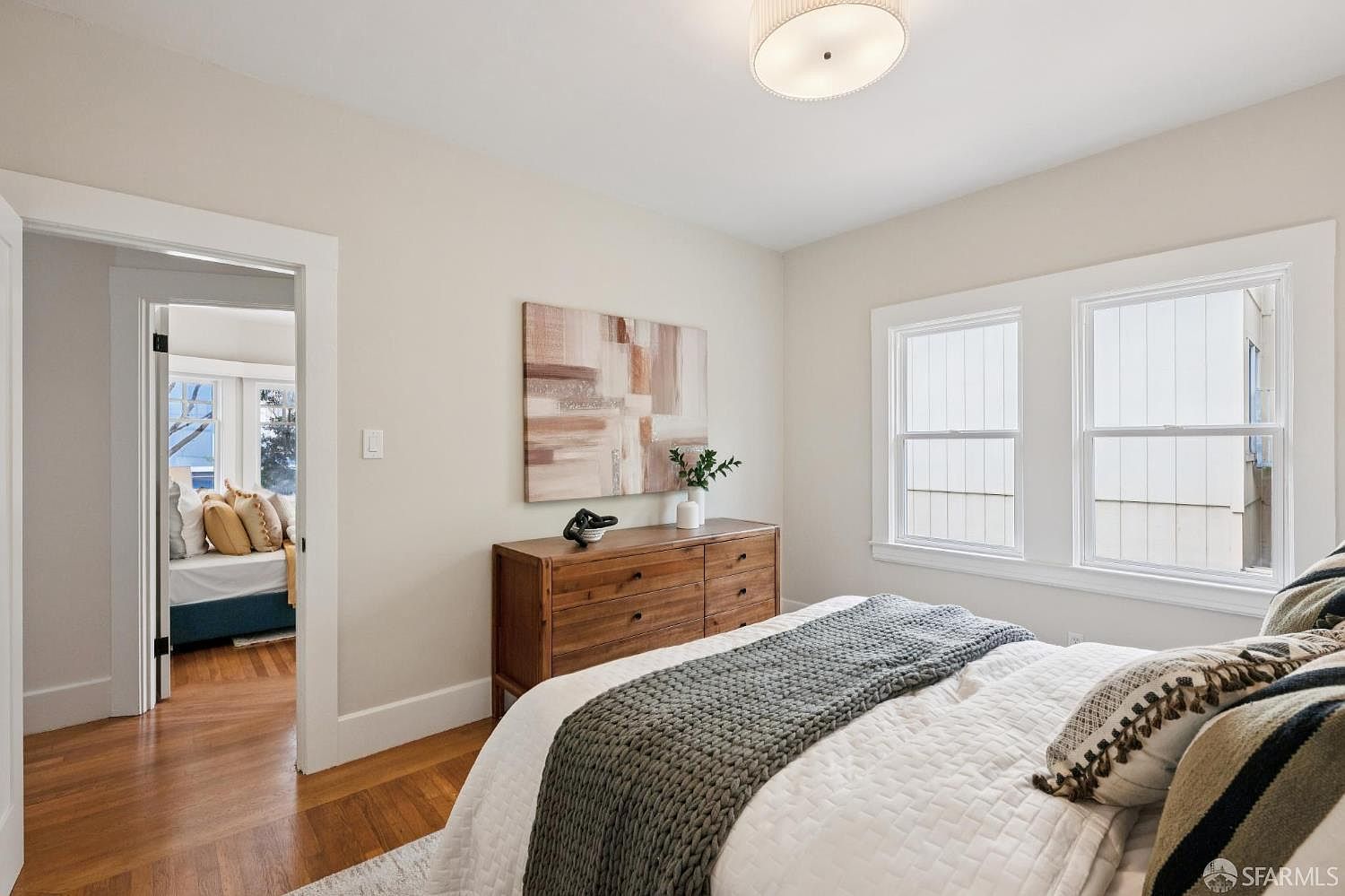 This bright and inviting bedroom features warm hardwood flooring and neutral wall tones, creating a cozy atmosphere. A wooden dresser sits against the wall beneath a piece of abstract art, while a bed with a textured gray throw blanket occupies the foreground. The room is illuminated by a central flush-mount light fixture and natural light streaming through large windows, with a doorway providing a glimpse into an adjacent bedroom.