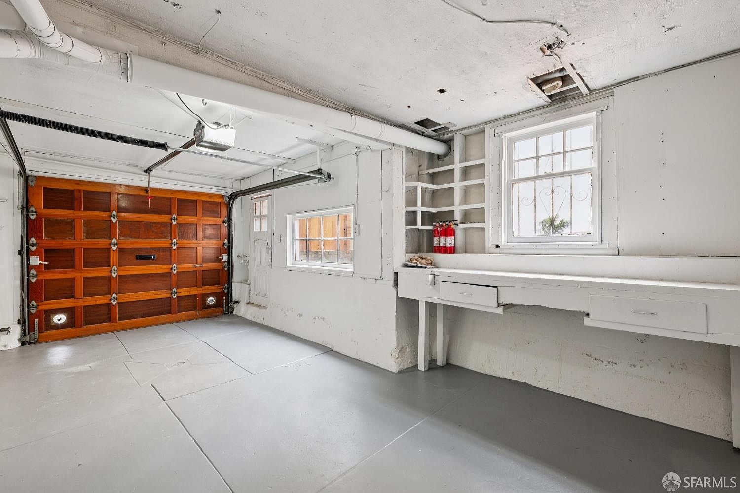 This image shows the interior of a functional garage space featuring a wooden sectional garage door and a painted concrete floor. To the right, a built-in workbench with drawers and open shelving provides storage, accompanied by a window that allows natural light into the area. The space is finished with white-painted walls and exposed overhead piping, presenting a clean and utilitarian environment.